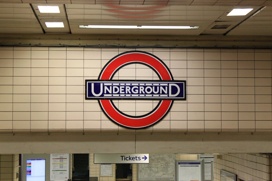 Close-up of a London Underground sign mounted on a light-colored brick wall, featuring a red circle with a horizontal blue bar across the middle displaying the word 'UNDERGROUND' in white text and a s