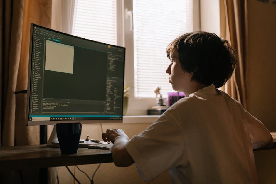 A person with short dark hair, dressed in a light-colored shirt, is seated at a wooden desk in a room with natural light coming through closed window blinds. They are looking at a large computer monit